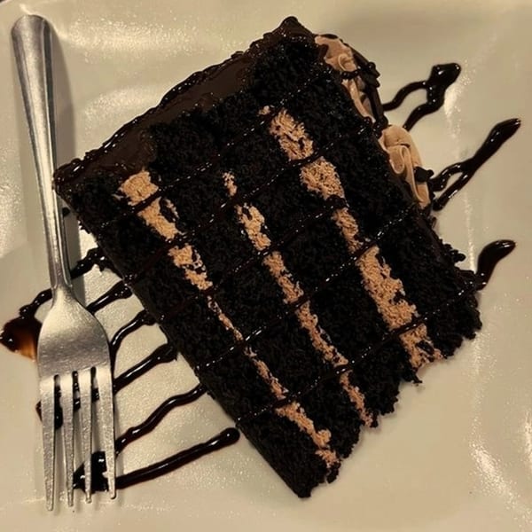 A slice of rich chocolate cake with light brown frosting layers and chocolate drizzle, next to a fork.
