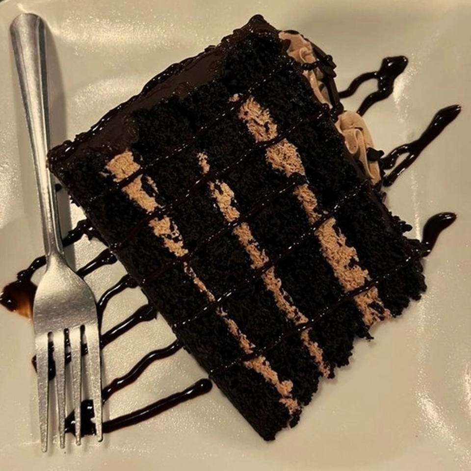 A slice of rich chocolate cake with light brown frosting layers and chocolate drizzle, next to a fork.