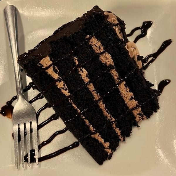 A slice of rich chocolate cake with mocha frosting layers and chocolate drizzle, served with a fork.