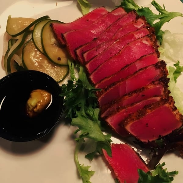Seared tuna slices with a dark crust, served with pickled cucumbers, greens, and a dipping sauce.