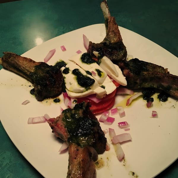Four lamb chops with pesto, mozzarella, tomatoes, and red onions on a white plate.